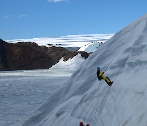 Two people scaling down an ice cliff with blue sky and rocky mountain in the background
