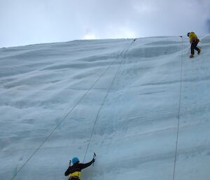 A person climbing an ice wall with an ice axe and crampons with a second person almost at the top