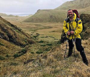 A man with walking poles smiles as he stands on a grassy hill