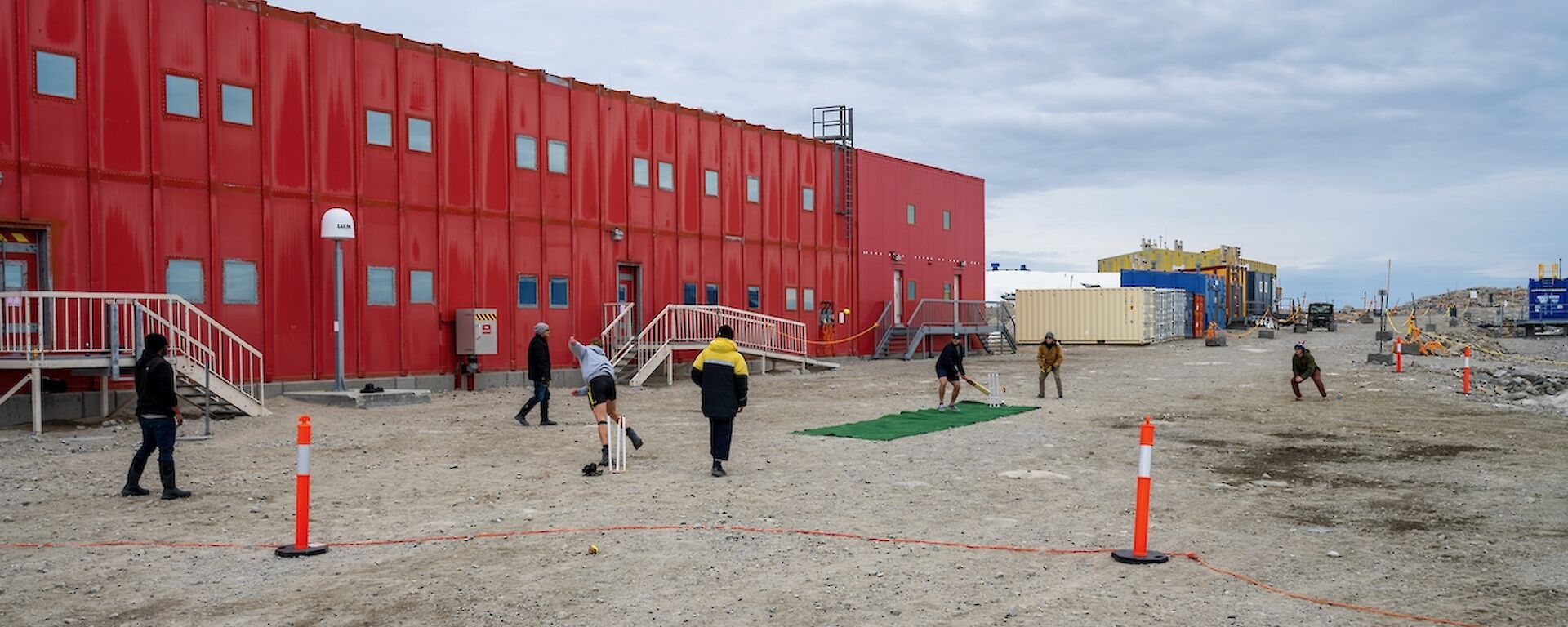 Scene of a cricket game in front of the Red Shed. A rectangle of green carpet has been laid out on the dirt road for a cricket pitch. The bowler has just thrown the ball and the batter is preparing to strike. Other fielders stand ready to catch the ball