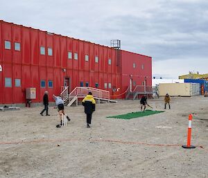 Scene of a cricket game in front of the Red Shed. A rectangle of green carpet has been laid out on the dirt road for a cricket pitch. The bowler has just thrown the ball and the batter is preparing to strike. Other fielders stand ready to catch the ball