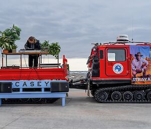 A DJ turntable and decorative pot plants in a flatbed hitched to a red Hägglunds vehicle. Two large speakers are arranged on a bench in front of the flatbed. A DJ is playing music from the turntable, leaning intently over the controls