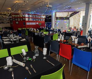 A large room arranged with dining tables and decorated with Christmas lights. The tables are covered with black cloth and laid with cutlery, napkins, glassware and Christmas crackers