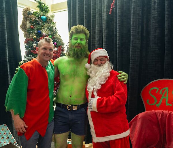 Three people, dressed up as a Christmas elf, Dr Seuss's Grinch and Santa Claus, huddled together and smiling for the camera