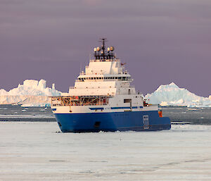 A blue ship sits in the sea surrounded by ice