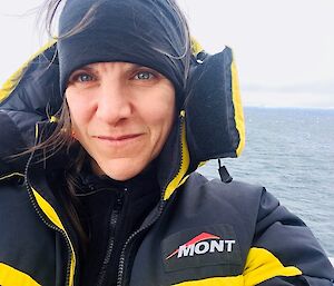 A woman in a warm jacket stares at the camera with the sea behind her