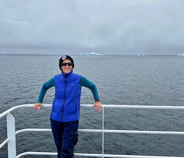 A woman smiling on board a ship with ice bergs behind her.