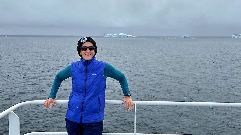 A woman smiling on board a ship with ice bergs behind her.