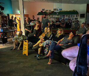 A roomful of people, some sitting on chairs, sofas or on the floor, some standing, looking up at a projector screen