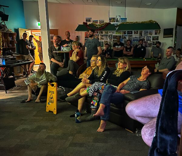 A roomful of people, some sitting on chairs, sofas or on the floor, some standing, looking up at a projector screen