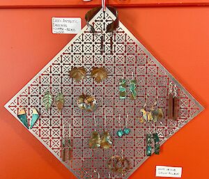 Handmade metal earrings of various designs, hung on a patterned, diamond-shaped metal screen which is tied to a wall fixture by a ribbon