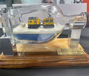 A miniature replica of a yellow Hägglunds vehicle on a fabricated bed of snow that has been constructed inside a horizontally-aligned glass bottle
