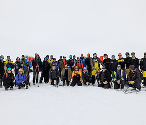 Thirty-six people in a snowy location, dressed in winter outdoors gear and arranged in two lines for a group photo. The people in the front row are crouching or kneeling in the snow. Some of the people are wearing or carrying skis and ski poles, and one of them is straddling a bicycle