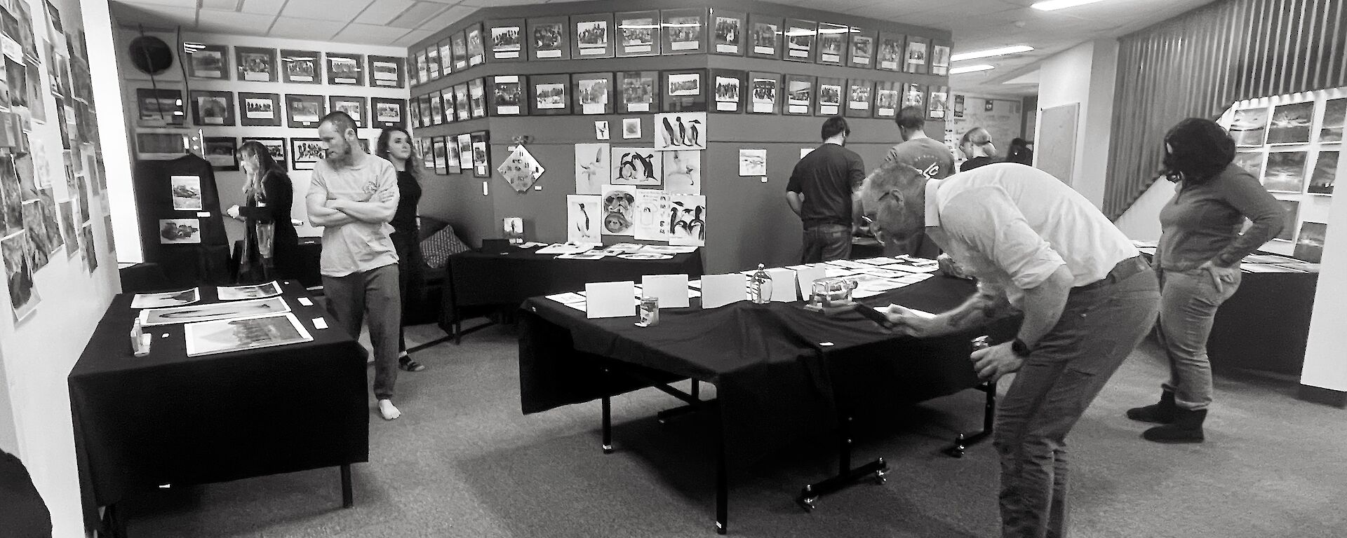 A scene of a room in which artwork and sculptures have been laid out on tables and hung on the walls. A number of people in the scene are closely examining various pieces of artwork