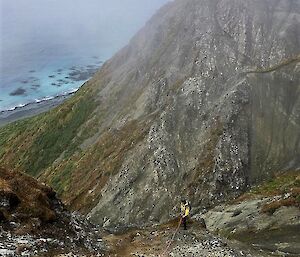 A man connected to a rope stands half way down a rocky hill