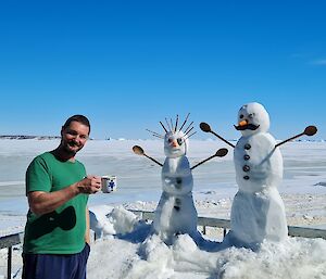 A man stands with a couple of snowmen holding his morning coffee