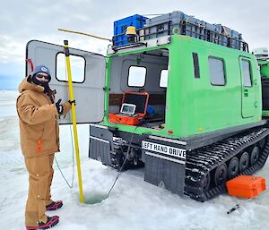 A man standing behind a Hagglunds vehicle holding a pole that leads into the sea ice