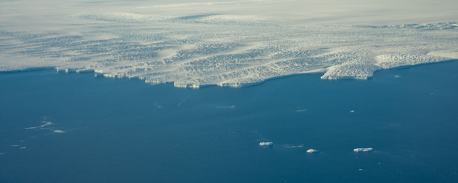 An aerial view of the edge of a glacier where it meets the ocean. The glacier looks like a great, white coastline, with low, dune-like wave formations and rugged, rock-like texture.
