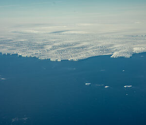 An aerial view of the edge of a glacier where it meets the ocean. The glacier looks like a great, white coastline, with low, dune-like wave formations and rugged, rock-like texture.