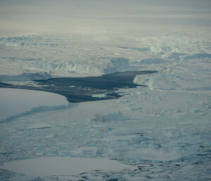 A view of ocean ice in many shapes and sizes. Some ice is in jagged chunks, some is shaped like small, flat plates. Large, thick ice sheets predominate the scene: some ocean water is visible, looking like a dark inlet between banks of ice.