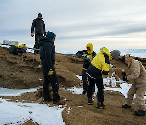 A team of people in outdoor gear standing on a gentle rock slope. A couple of people are holding a metal pole horizontal and steady, while another person mounts a small anemometer on the end of the pole.