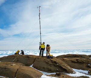 The metal pole with anemometer attached has been fixed upright on top of the rocks with guy wires. Two people to the left are securing one of the guy wires, whilst another two inspect the base of the pole.