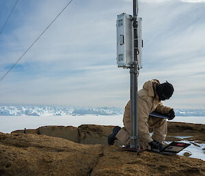 A person in thick outdoor work clothing, a balaclava and gloves is kneeling at the base of the pole and looking down at a laptop computer.