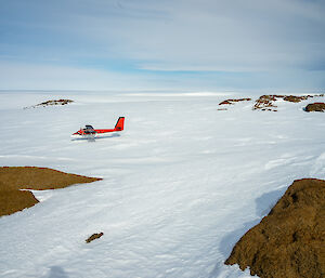 A small Twin Otter plane, coloured red on top and white underneath, stands out against the snow it is parked on. Some rocks break up through the snow around the edges of the scene