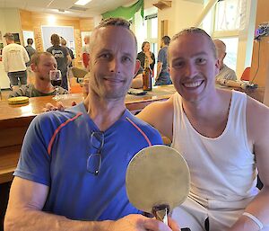 A photo of two men in casual sports attire seated next to each other at a bar counter, smiling for the camera. The man on the left is holding a Ping-Pong paddle inscribed with the words: "Balls of Fury Champion - 75th ANARE Casey"