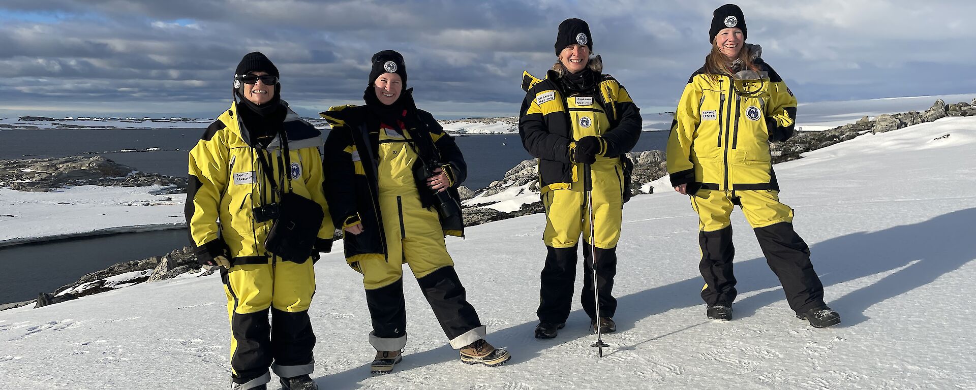 Four women wearing yellow and black Antarctic weather-proof clothing, standing in a row and smiling for the camera. The ground is covered with snow, behind them is a rocky coastline and a view of an ocean bay