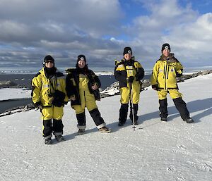 Four women wearing yellow and black Antarctic weather-proof clothing, standing in a row and smiling for the camera. The ground is covered with snow, behind them is a rocky coastline and a view of an ocean bay
