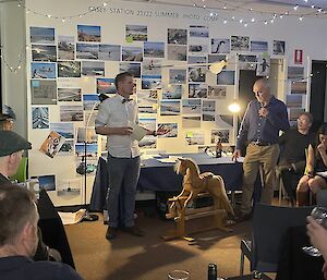 The photo centres on a wooden rocking horse. The maker of the rocking horse stands to the right, looking down at his creation. To the left of the rocking-horse stands an auctioneer, speaking to a roomful of seated people, a few of whom can be seen at the edges of the photo