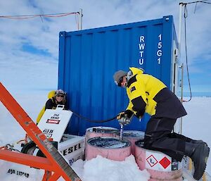 2 expeditioners filling up a generator with fuel from pink fuel drums next to a blue shipping container