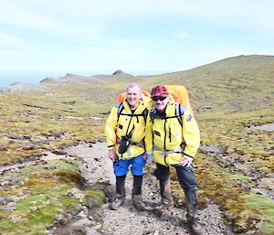 Two men with yellow jackets and backpacks stand on a walking track