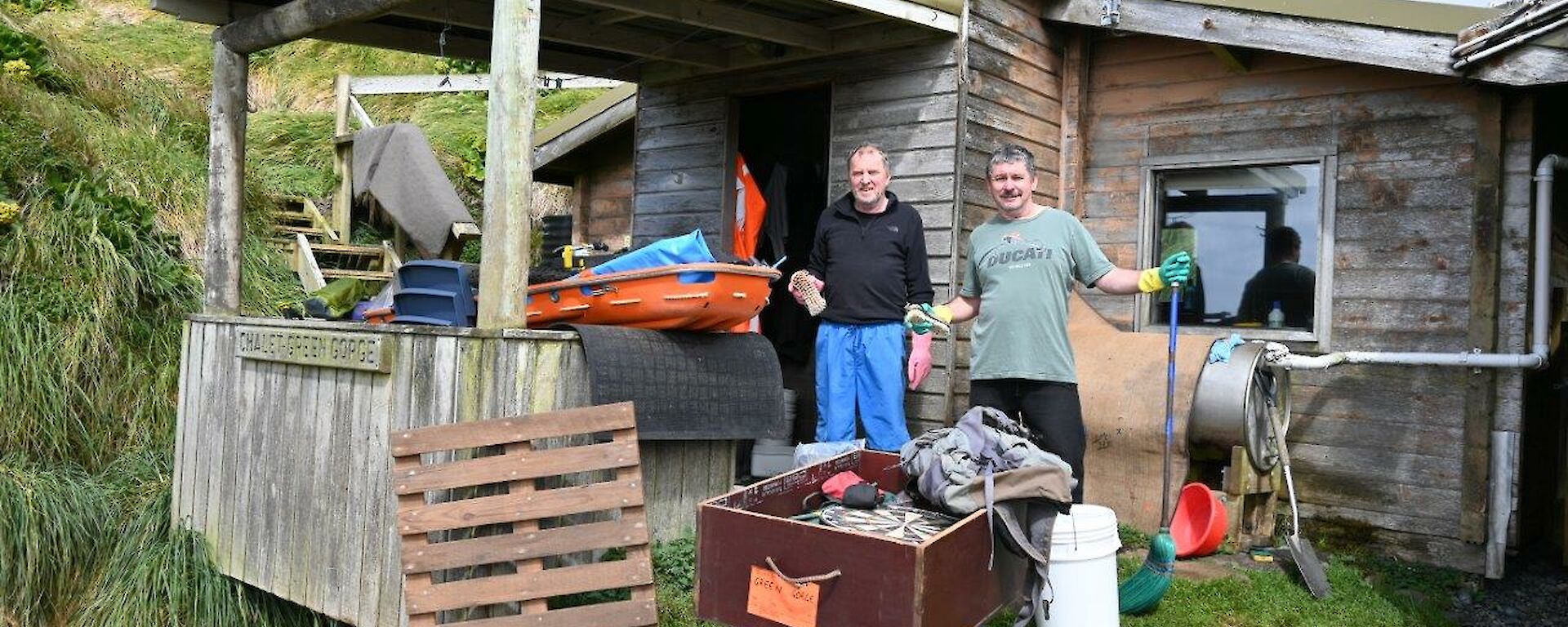 Two men stand outside a wooden hut as they are cleaning it