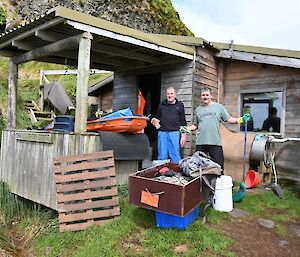 Two men stand outside a wooden hut as they are cleaning it