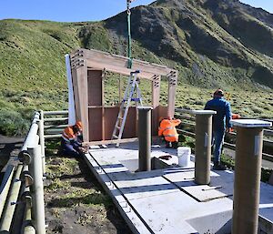 Three people work on a small building in front of a large grassy hill