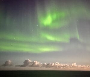The ocean reflects the green of the aurora as white clouds hover on the horizon