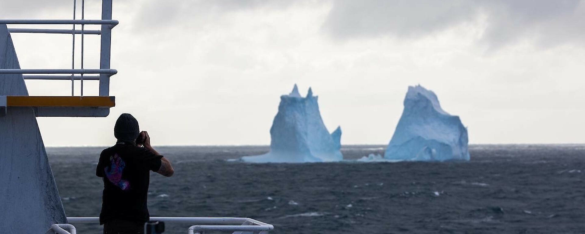 Two icebergs sit in a dark sea as a photographer stands on the front of the ship taking photos