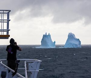 Two icebergs sit in a dark sea as a photographer stands on the front of the ship taking photos