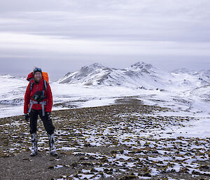 A hiker in red stands on a ridge with snow covered hills all around
