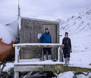 Two people stand on the wooden deck of a hut on a snowy hillside