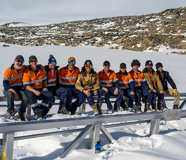 Ten tradesmen wearing high-vis work clothing, sunglasses, caps and beanies seated together in a row on a metal beam and smiling for the camera. The ground is covered with snow and a low, rocky hillside can be seen behind them