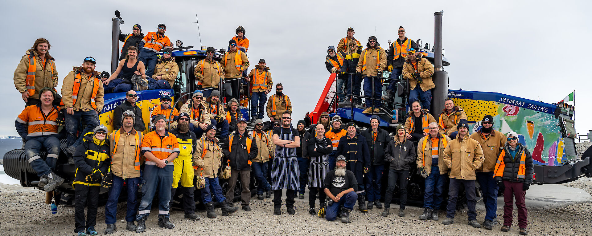 A group of about 40 people posing for a team photo. Many are wearing tan jackets or other cold weather gear, a couple in the front are wearing chef's aprons. Most are standing or sitting on two brightly-painted Challenger tractors, with one row of people standing in front