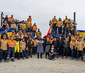 A group of about 40 people posing for a team photo. Many are wearing tan jackets or other cold weather gear, a couple in the front are wearing chef's aprons. Most are standing or sitting on two brightly-painted Challenger tractors, with one row of people standing in front