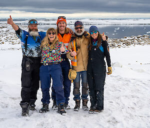 A team of five people posing for the camera with smiles and thumbs-up gestures, a couple of them are wearing colourful shirts. They are standing on snowy ground in front of a calm bay under a cloudy sky