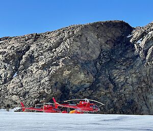 Two red helicopters on the ice in front of some rocky hills