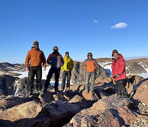 Five people standing on rocks with snow and blue sky in the background