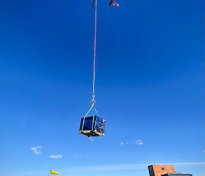 A helicopter carrying a blue cage on a long rope