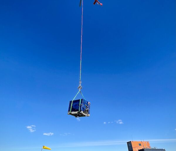 A helicopter carrying a blue cage on a long rope